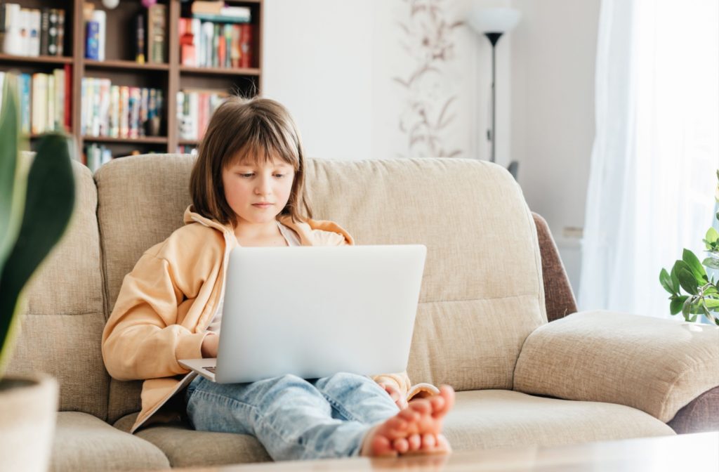 A young girl sitting on the couch doing schoolwork on a laptop.