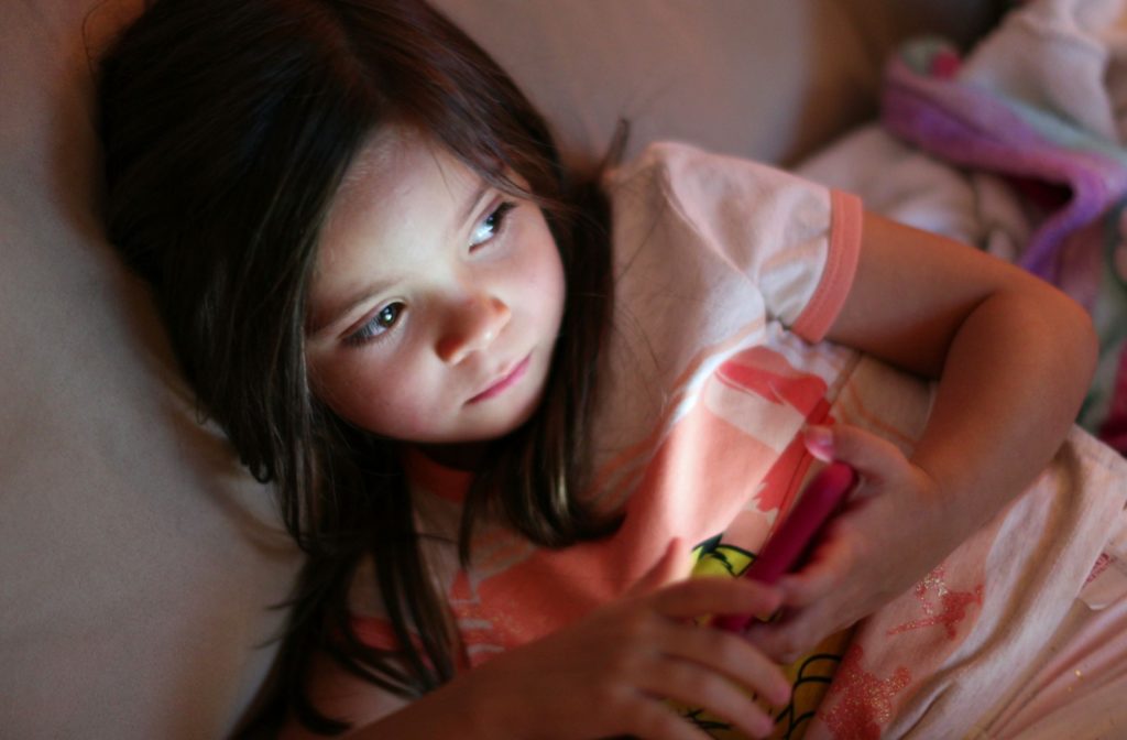 A young girl laying in bed at night with a smartphone in her hand, staring at a tv screen.
