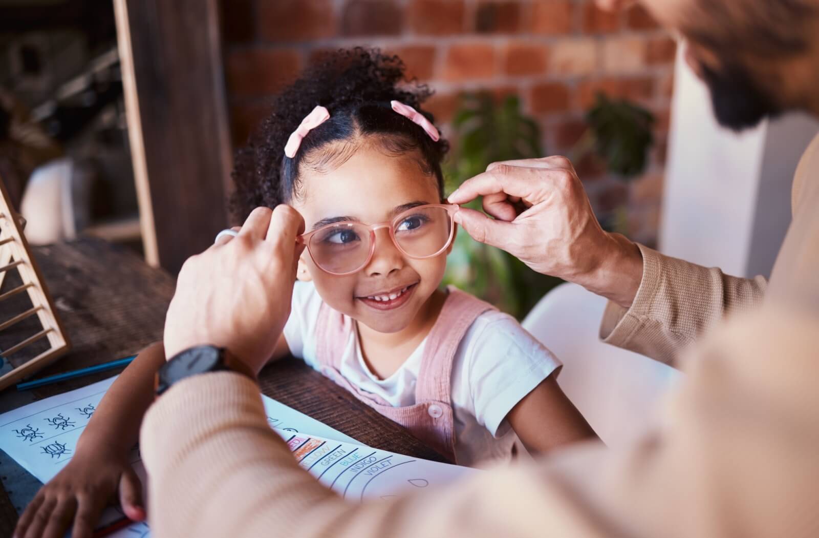 Smiling young girl trying on new glasses with help from an adult during an eye exam or fitting.